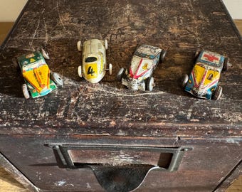 May include: Four vintage toy race cars in various colors, including yellow, green, red, and blue. The cars are made of metal and have a distressed appearance. They are arranged on a dark wooden surface, with a white ceramic object in the background.