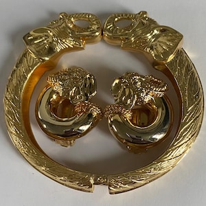 May include: Gold-toned jewelry set featuring a bracelet and a pair of clip-on earrings. The bracelet has two elephant head designs on each end, and the earrings have elephant head designs. The bracelet and earrings have a shiny, polished finish.