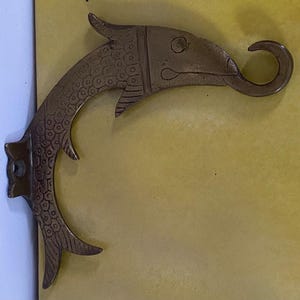 May include: A brass-colored, fish-shaped wall hook. The hook features a curved body with a fish head and tail design. The fish's body has a scale pattern, and the hook has a mounting plate with a hole for hanging.