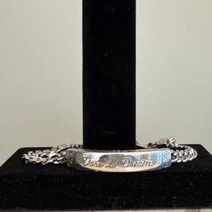 K&ouml;nnte beinhalten: Ein silbernes Armband mit einem rechteckigen Mittelst&uuml;ck, das mit den Worten "Dare to Dream" in Schreibschrift graviert ist. Das Armband hat ein Panzerketten-Design und wird auf einem schwarzen St&auml;nder pr&auml;sentiert.