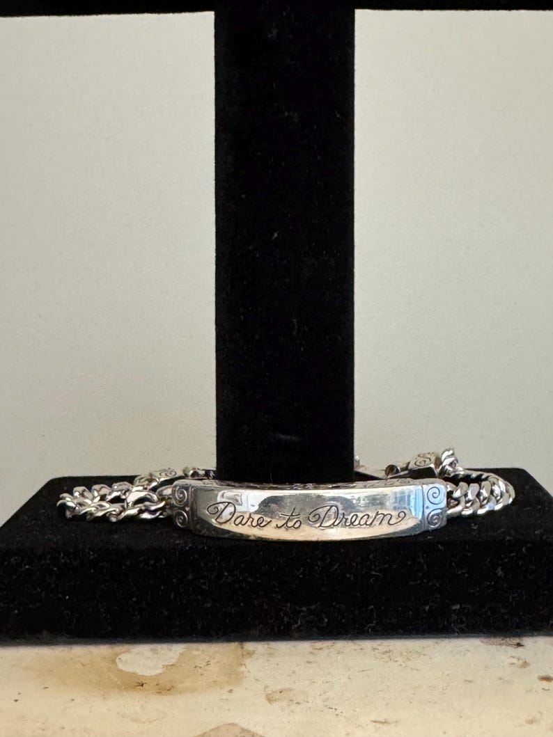 K&ouml;nnte beinhalten: Ein silbernes Armband mit einem rechteckigen Mittelst&uuml;ck, das mit den Worten "Dare to Dream" in Schreibschrift graviert ist. Das Armband hat ein Panzerketten-Design und wird auf einem schwarzen St&auml;nder pr&auml;sentiert.