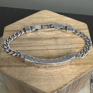 K&ouml;nnte beinhalten: Ein silberfarbenes Armband mit einem Panzerketten-Design. Das Armband hat ein rechteckiges Mittelteil mit der Gravur "Dare to Dream" und dekorativen Wirbeln. Der Verschluss ist im Faltstil gehalten.