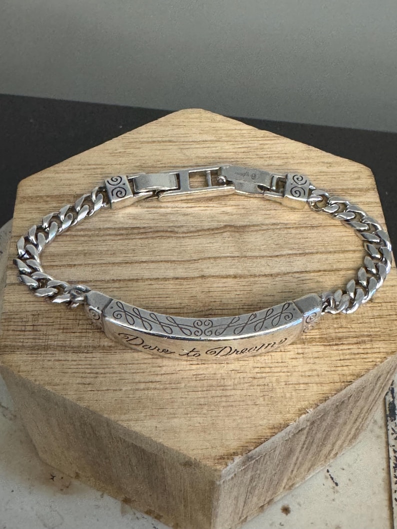 K&ouml;nnte beinhalten: Ein silberfarbenes Armband mit einem Panzerketten-Design. Das Armband hat ein rechteckiges Mittelteil mit der Gravur "Dare to Dream" und dekorativen Wirbeln. Der Verschluss ist im Faltstil gehalten.
