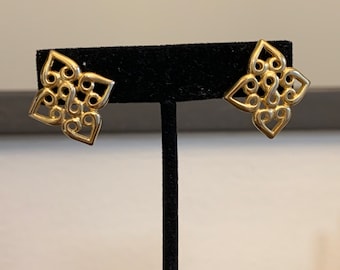 May include: A pair of gold-toned earrings with a geometric, intricate design. The earrings are displayed on a black stand.