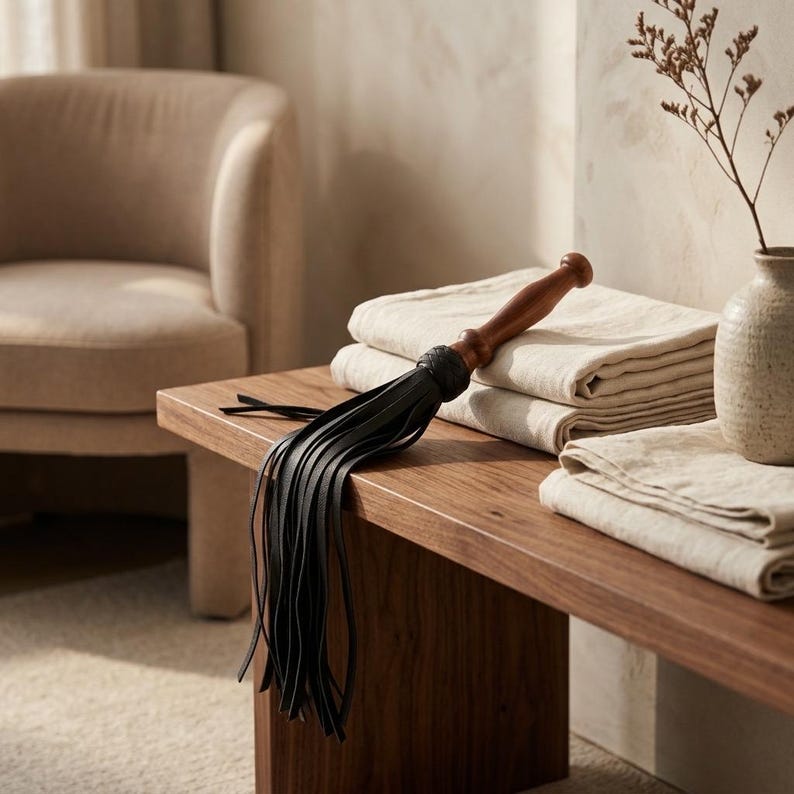 May include: A black leather flogger featuring a smooth, dark brown wooden handle with a braided leather collar. The flogger consists of multiple long, thin black leather strips attached to the handle, resting on a stack of folded beige linen fabric.