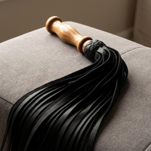May include: A flogger featuring a smooth, polished wooden handle with a rounded end. The handle connects to a braided leather collar that transitions into numerous long, thin, black leather strips that drape downward in a cascading arrangement.