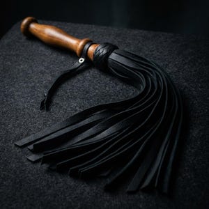 May include: A flogger featuring a smooth, polished brown wooden handle with a rounded pommel. A thin leather strap with a metal eyelet is attached to the handle. The head consists of numerous long, black leather strips secured by a braided knot.