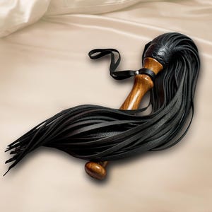 May include: A multi-strand flogger featuring a polished wooden handle with a smooth, contoured grip. The top of the handle is wrapped in black textured material, from which numerous long, thin black leather strips extend outward in a fan shape.