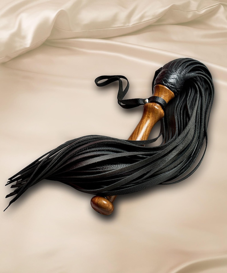 May include: A multi-strand flogger featuring a polished wooden handle with a smooth, contoured grip. The top of the handle is wrapped in black textured material, from which numerous long, thin black leather strips extend outward in a fan shape.