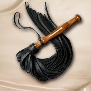 May include: A flogger featuring a polished brown wooden handle and a braided black leather collar. Attached to the handle are numerous long, thin black leather strips that fan out in a circular pattern, accompanied by a thin black leather wrist loop.