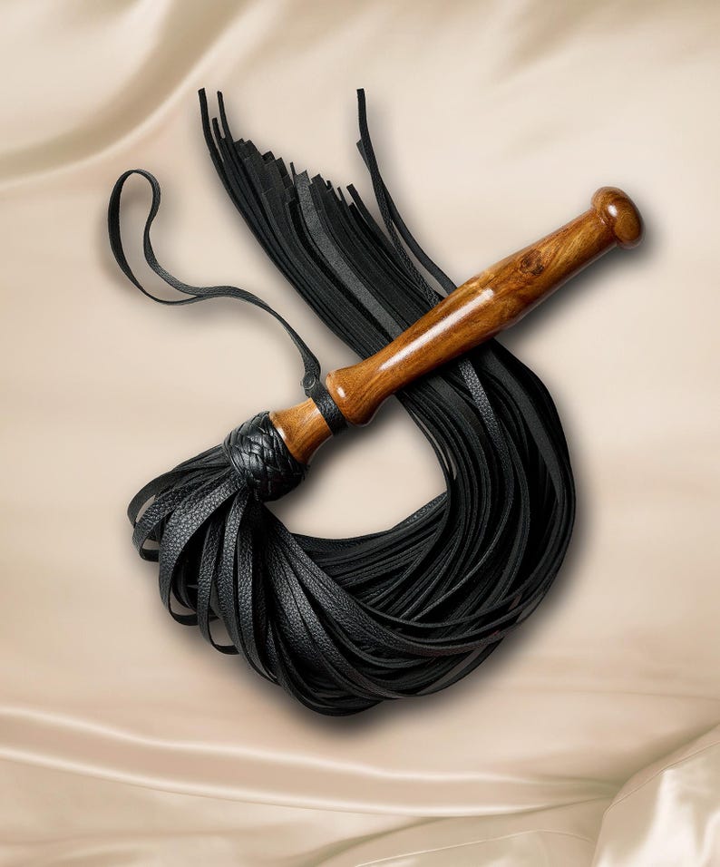 May include: A flogger featuring a polished brown wooden handle and a braided black leather collar. Attached to the handle are numerous long, thin black leather strips that fan out in a circular pattern, accompanied by a thin black leather wrist loop.