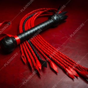 May include: A black leather whip with a red braided handle and multiple red braided tails. The whip is lying on a red surface.