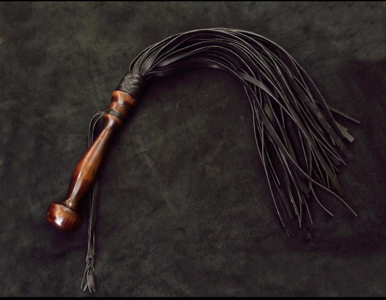 May include: A multi-tailed whip featuring a polished dark brown wooden handle with a rounded pommel and a braided leather collar. Attached to the handle are numerous long, thin, flexible black leather strips that fan out in a curved arc.