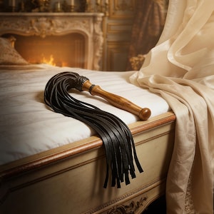 May include: A multi-tailed whip featuring a smooth, polished wooden handle with a rounded pommel and a series of long, flat black leather strips attached to the top. The leather strands are secured with a tightly wrapped black leather cord.