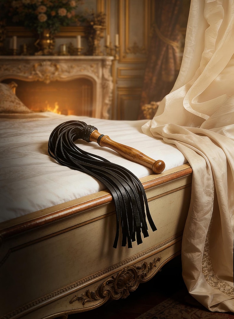 May include: A multi-tailed whip featuring a smooth, polished wooden handle with a rounded pommel and a series of long, flat black leather strips attached to the top. The leather strands are secured with a tightly wrapped black leather cord.