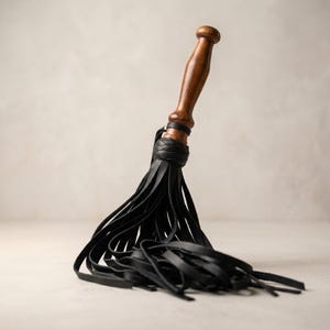 May include: A multi-tailed whip featuring a smooth, polished brown wooden handle with a rounded pommel. The handle connects to a cluster of numerous thin, flat black leather strips that fan out and drape downwards in a dense, layered arrangement.