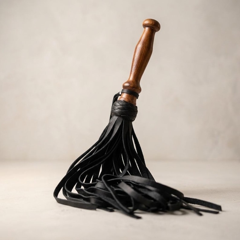 May include: A multi-tailed whip featuring a smooth, polished brown wooden handle with a rounded pommel. The handle connects to a cluster of numerous thin, flat black leather strips that fan out and drape downwards in a dense, layered arrangement.