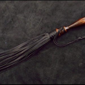 May include: A flogger featuring a polished dark brown wooden handle with a rounded pommel and a series of long, thin black leather strips attached to the base of the handle with a black leather wrap and a thin black leather cord tie.