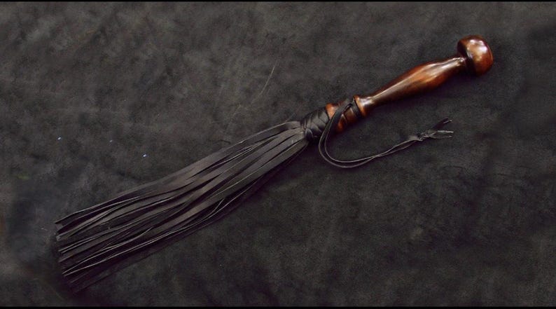 May include: A flogger featuring a polished dark brown wooden handle with a rounded pommel and a series of long, thin black leather strips attached to the base of the handle with a black leather wrap and a thin black leather cord tie.