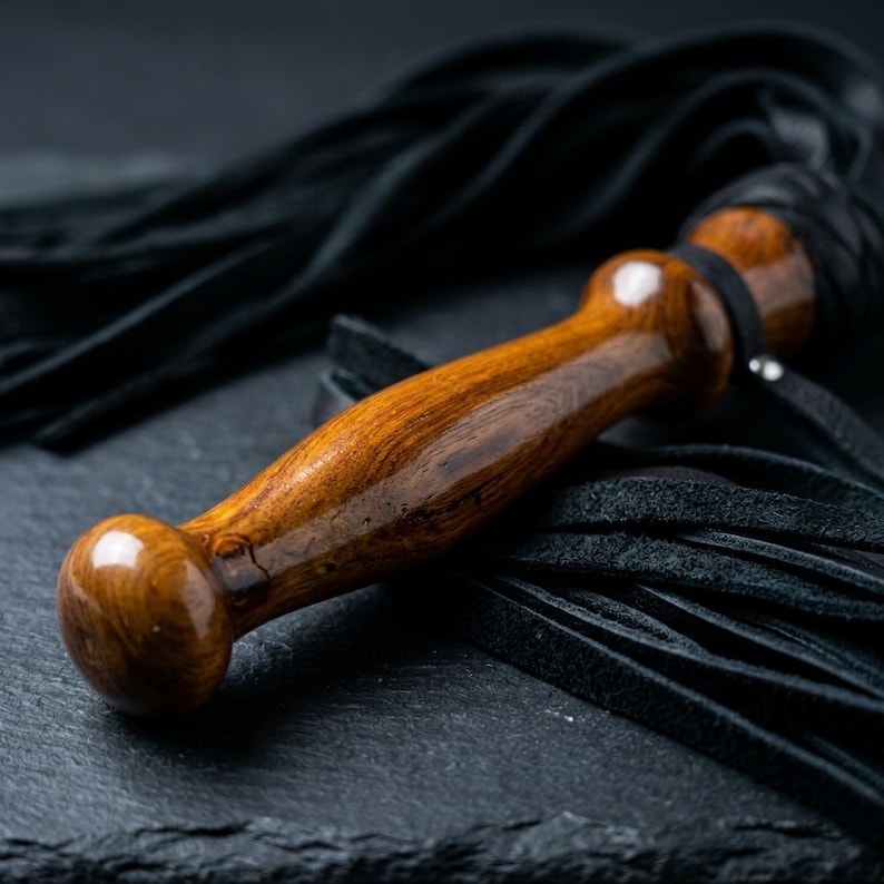 May include: A whip featuring a polished, smooth, brown wooden handle with a rounded pommel and a flared grip. Attached to the handle are numerous long, thin, black leather strips that extend outward, creating a dense, textured fringe effect.