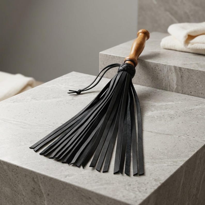 May include: A flogger featuring a smooth, light brown wooden handle and a braided collar connecting to numerous long, thin, black leather strips. The handle has a rounded knob at the end, and the black leather tails extend outward in a fan shape.