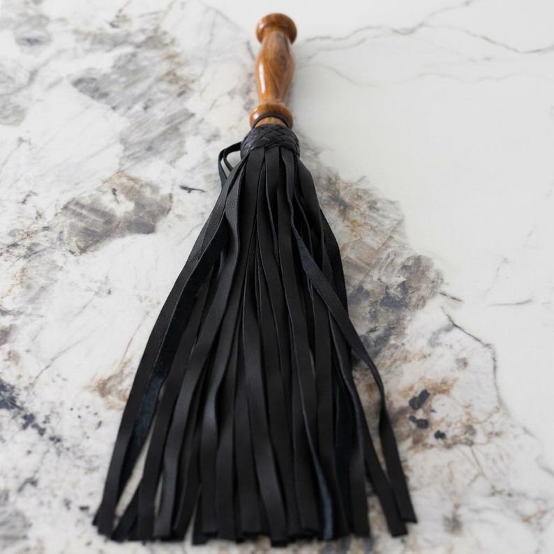 May include: A flogger featuring a polished brown wooden handle with a rounded pommel and a braided collar, attached to a cluster of numerous long, thin, black leather strips that hang downward in a dense, uniform arrangement.