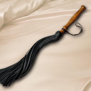 May include: A whip featuring a polished brown wooden handle with a flared end and a black leather wrist strap. The handle connects to a cluster of long, thin black leather strips that are secured by a braided black leather knot at the base.