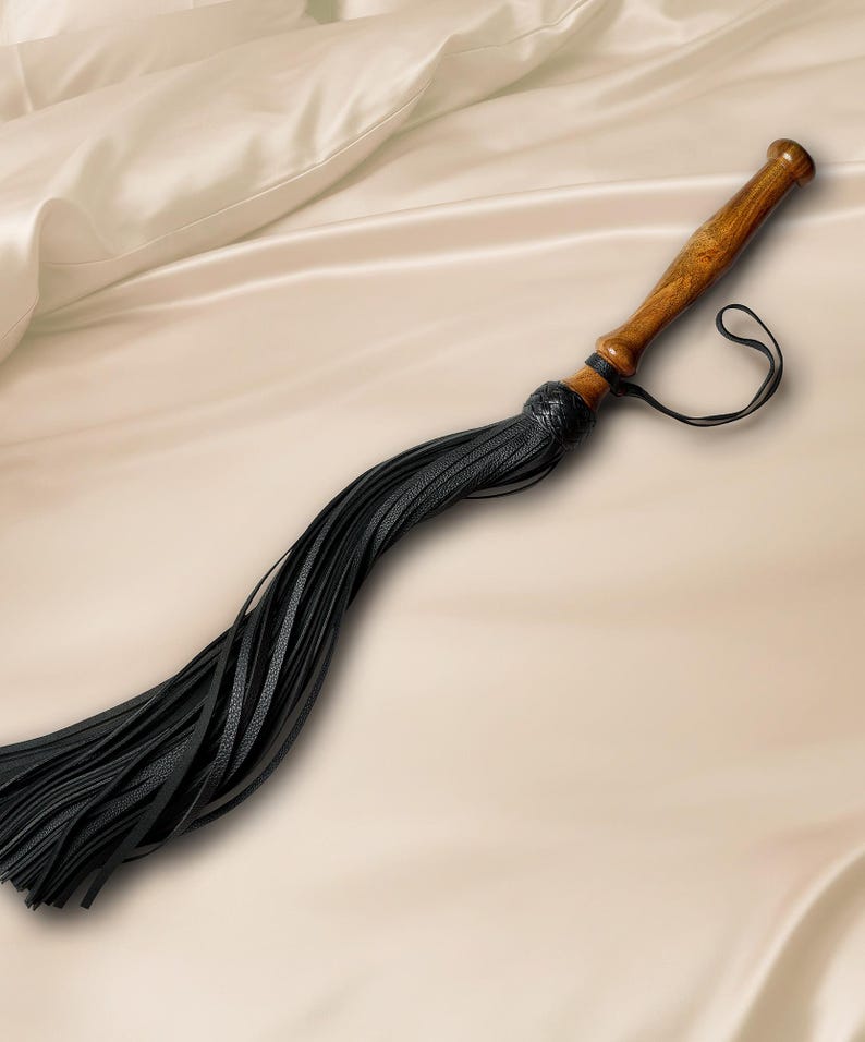 May include: A whip featuring a polished brown wooden handle with a flared end and a black leather wrist strap. The handle connects to a cluster of long, thin black leather strips that are secured by a braided black leather knot at the base.