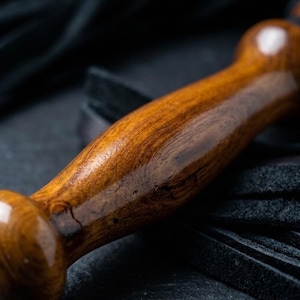 May include: A polished, cylindrical wooden handle with a rich brown grain and a smooth, glossy finish. The handle features a rounded knob at the end and is attached to a cluster of black leather strips that extend from the base of the wood.