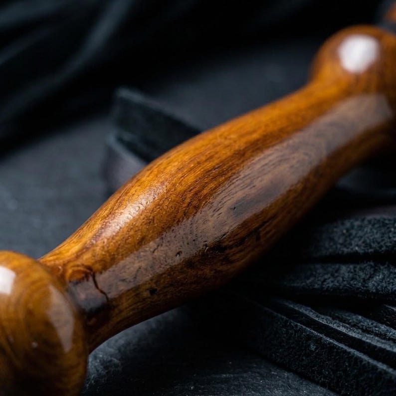 May include: A polished, cylindrical wooden handle with a rich brown grain and a smooth, glossy finish. The handle features a rounded knob at the end and is attached to a cluster of black leather strips that extend from the base of the wood.