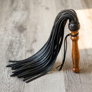 May include: A black leather flogger featuring a polished wooden handle with a natural grain pattern. The handle is topped with a braided black leather knot where multiple long, thin black leather strips are attached, creating a cascading effect.
