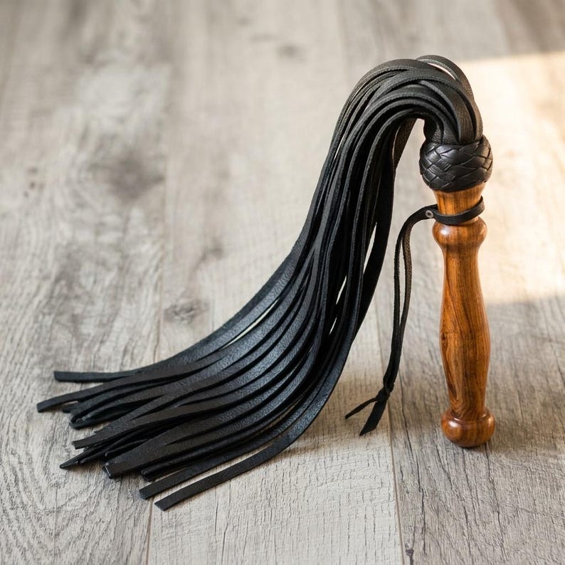 May include: A black leather flogger featuring a polished wooden handle with a natural grain pattern. The handle is topped with a braided black leather knot where multiple long, thin black leather strips are attached, creating a cascading effect.