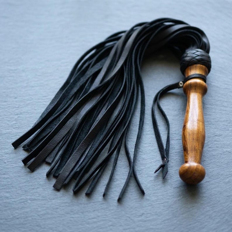 May include: A multi-tailed whip featuring a polished, light brown wooden handle with a rounded pommel. A braided black leather collar connects the handle to numerous long, thin, flexible black leather strips that fan out from the base.