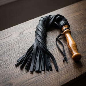 May include: A black leather flogger featuring multiple long, thin strips attached to a polished, light brown wooden handle. The handle has a smooth, contoured shape with a braided leather knot at the top and a thin leather wrist strap attached.