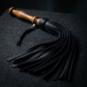 May include: A flogger featuring a smooth, polished brown wooden handle with a rounded pommel. A black leather strap with a silver-toned metal eyelet is attached to the handle. The head consists of multiple long, black leather strips.