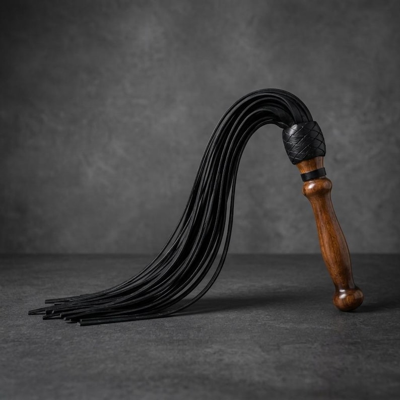 May include: A multi-tailed whip featuring a polished brown wooden handle with a rounded pommel. The top of the handle is wrapped in a black braided leather collar, from which numerous long, thin, flexible black leather strips extend downward.
