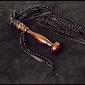 May include: A multi-tailed whip featuring a polished dark brown wooden handle with a rounded pommel and a braided leather collar. Numerous long, thin black leather strips are attached to the handle, accompanied by a thin black leather wrist strap.