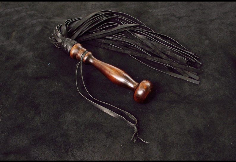 May include: A multi-tailed whip featuring a polished dark brown wooden handle with a rounded pommel and a braided leather collar. Numerous long, thin black leather strips are attached to the handle, accompanied by a thin black leather wrist strap.