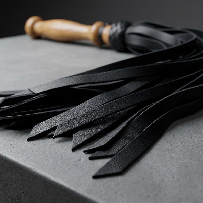 May include: A flogger featuring a smooth, light-toned wooden handle connected to a cluster of long, black leather strips. The base of the handle is secured with a braided black leather knot, transitioning into multiple flat, flexible black tails.