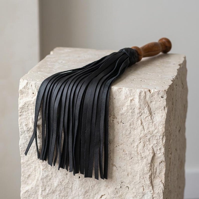 May include: A black leather flogger featuring a series of long, thin, flexible strips attached to a smooth, rounded wooden handle. The handle is carved with a bulbous end, and the black leather tails are secured at the top with a braided knot.