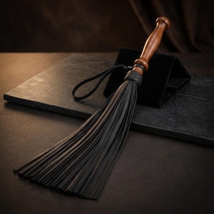 May include: A flogger featuring a polished brown wooden handle with a rounded pommel, a black braided leather collar, and a long black leather wrist strap. The head consists of numerous thin, flexible black leather strips hanging downward.