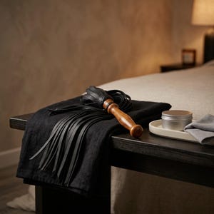 May include: A black leather flogger with a polished wooden handle featuring multiple long, thin leather strips attached to a braided leather collar. The flogger rests on a folded black fabric cloth next to a small silver metal tin container.