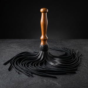 May include: A multi-tailed whip featuring a smooth, polished brown wooden handle with a rounded top. The base of the handle is secured with a braided black leather collar, from which numerous thin, flexible black leather strips extend outward.