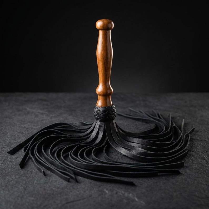May include: A multi-tailed whip featuring a smooth, polished brown wooden handle with a rounded top. The base of the handle is secured with a braided black leather collar, from which numerous thin, flexible black leather strips extend outward.