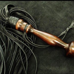 May include: A multi-strand black leather flogger featuring a polished dark brown wooden handle with a rounded pommel. The leather strips are attached to the handle with a braided black leather collar and a thin leather tie is attached to the grip.