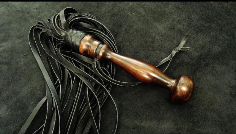 May include: A multi-strand black leather flogger featuring a polished dark brown wooden handle with a rounded pommel. The leather strips are attached to the handle with a braided black leather collar and a thin leather tie is attached to the grip.