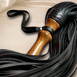 May include: A multi-tailed whip featuring a polished wooden handle with a smooth, contoured grip. The top of the handle is adorned with a black leather braided knot, which connects to numerous long, thin black leather strips and a wrist strap.