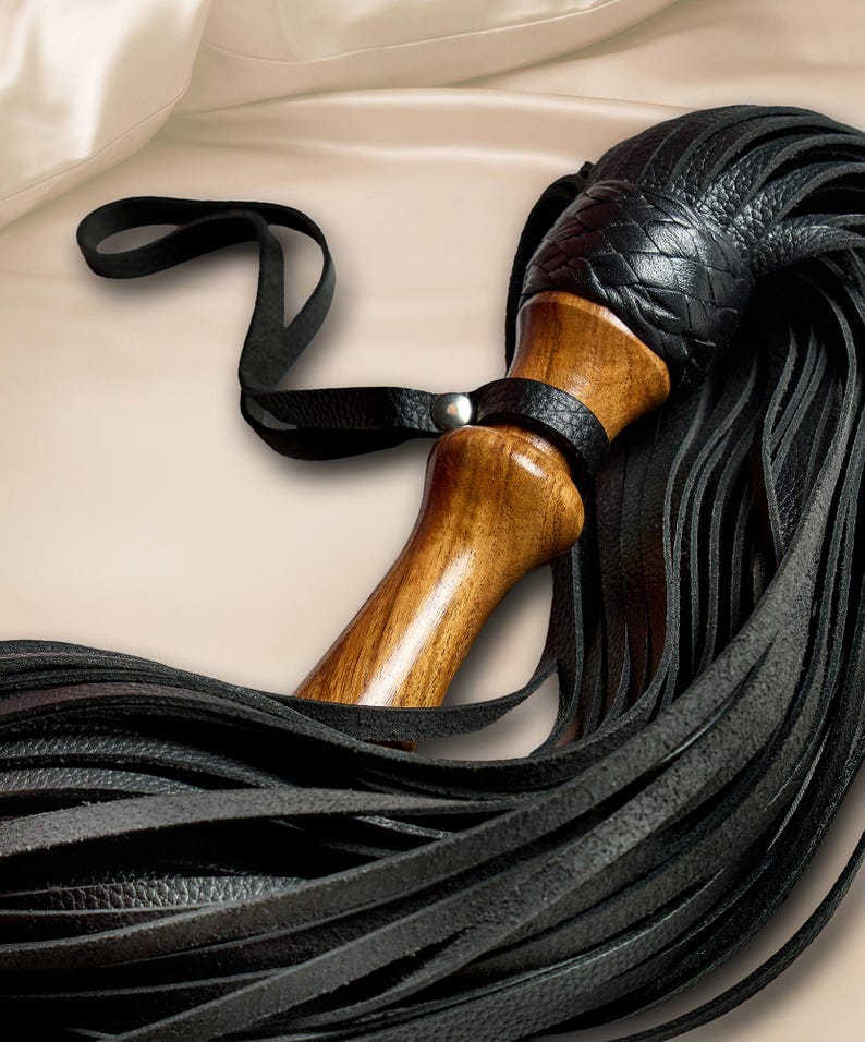 May include: A multi-tailed whip featuring a polished wooden handle with a smooth, contoured grip. The top of the handle is adorned with a black leather braided knot, which connects to numerous long, thin black leather strips and a wrist strap.