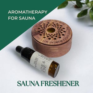 May include: Aromatherapy for sauna set. The image shows a wooden sauna diffuser with a patterned top and a small glass bottle of sauna freshener. The bottle has a black cap and a label that reads "SAUNA FRESHENER". The text "AROMATHERAPY FOR SAUNA" is also visible.