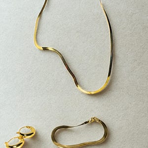 May include: A gold-colored jewelry set featuring a herringbone chain necklace, a bracelet, and a pair of hoop earrings. The necklace and bracelet have a flat, flexible design. The earrings are small, rounded hoops. The set is displayed on a light gray surface.